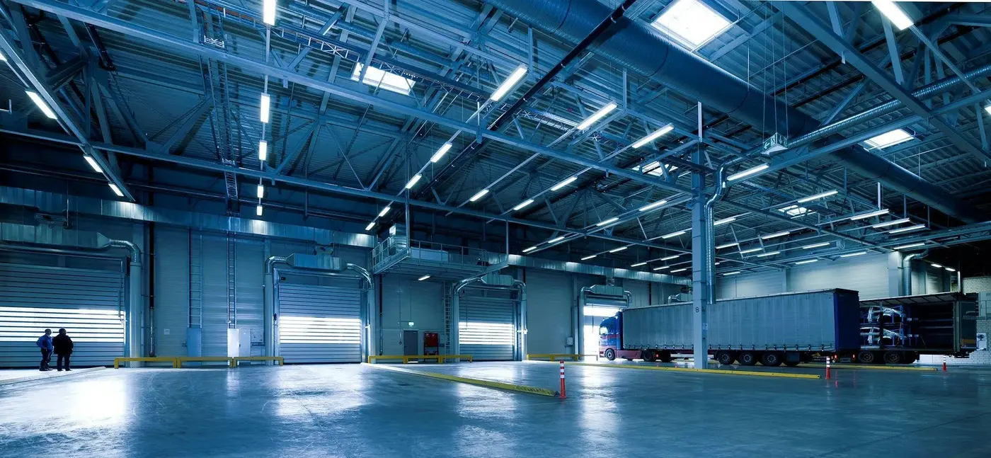 Inside a distribution warehouse: steel roof joists, ductwork and rows of high-bay lights above the dock doors, with a trailer backed in at a loading bay